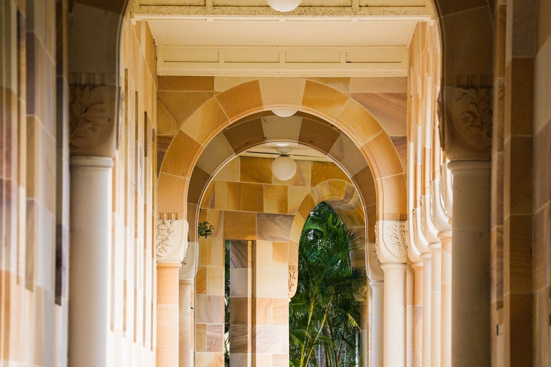 A sandstone cloister of round arches and columns, with a wooden bench along the wall and palms beyond the far archway
